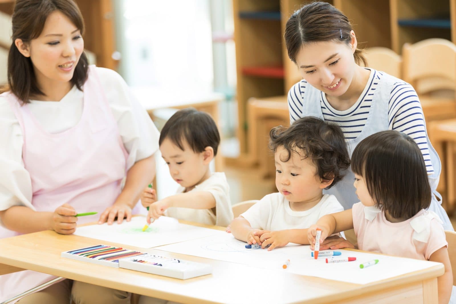 保育室の机で、お絵かきをする3人の園児に付き添う2人の保育士